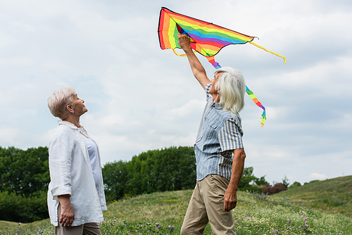 happy senior couple in casual clothes looking at flying kite and standing on green hill