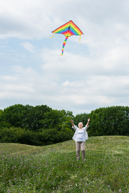 full length of senior woman in casual clothes looking at flying kite in cloudy sky