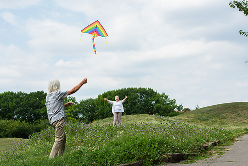 happy senior woman in casual clothes looking at husband playing with kite on green hill