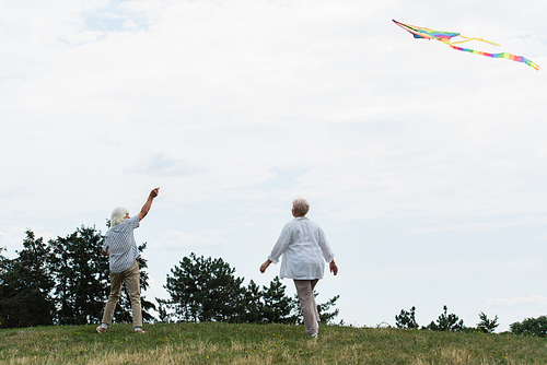 senior woman in casual clothes walking near husband playing with kite on green hill