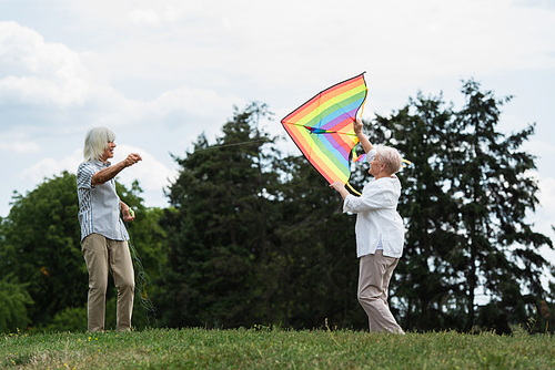 senior woman in casual clothes playing with kite near happy husband on green hill