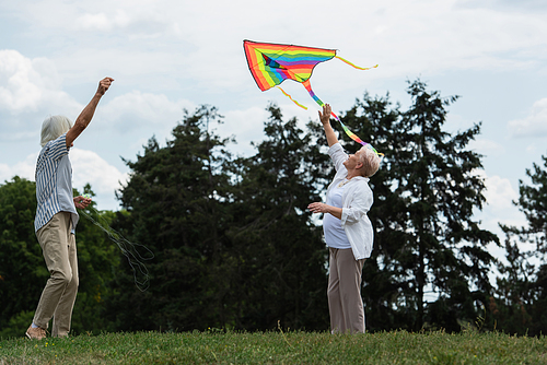 happy senior woman in casual clothes playing with kite near husband on green hill
