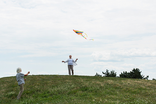 cheerful senior woman in casual clothes looking at kite near husband on green hill