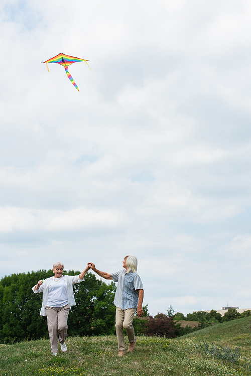 happy senior couple in casual clothes holding hands while playing with kite on green hill