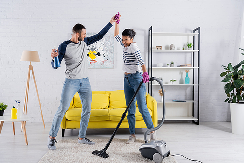 Happy multiethnic couple with cleaning supplies dancing in living room