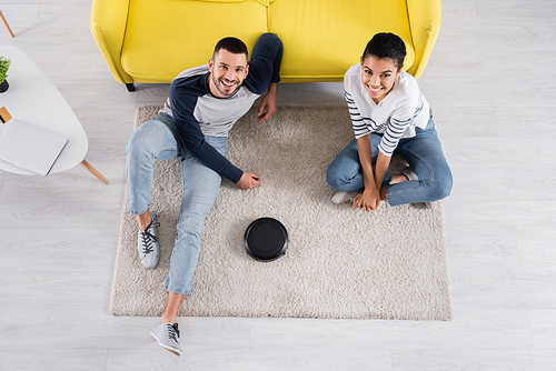 Overhead view of smiling interracial couple looking at camera near robotic vacuum cleaner on carpet