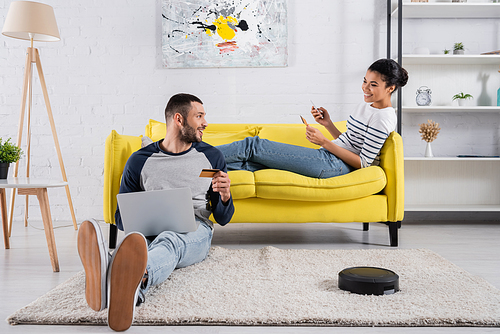 Interracial couple with gadgets and credit cards near robotic vacuum cleaner at home