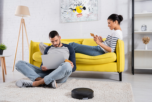 Smiling multiethnic couple using devices and credit cards near robotic vacuum cleaner