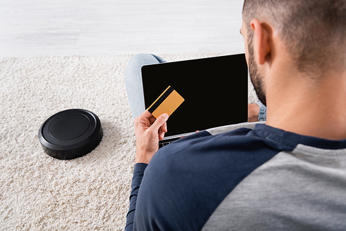 Back view of blurred man holding credit card and laptop near robotic vacuum cleaner