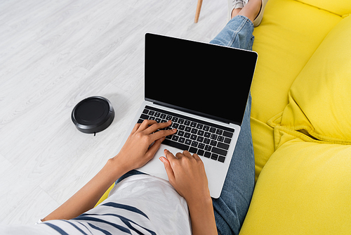 Cropped view of african american woman using laptop with blank screen near robotic vacuum cleaner