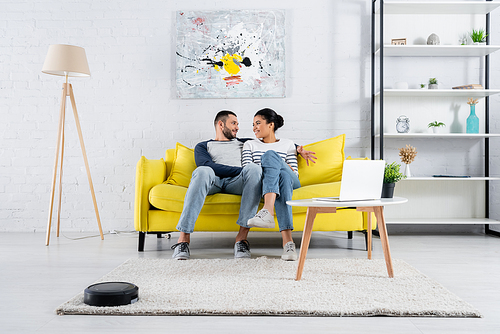 Smiling interracial couple talking near laptop and robotic vacuum cleaner