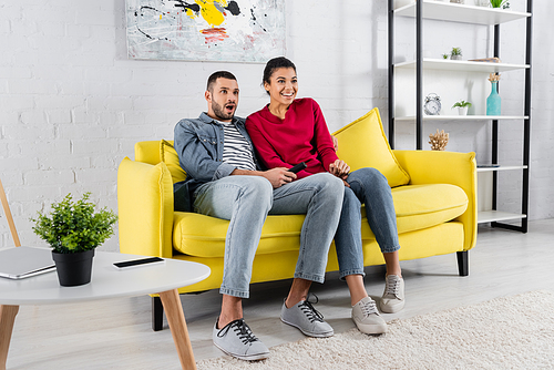 Smiling african american woman sitting near amazed boyfriend with remote controller on couch