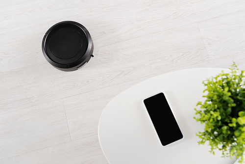 Top view of robotic vacuum cleaner on floor near smartphone on coffee table
