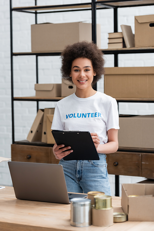 happy african american woman standing with clipboard near laptop in volunteer center