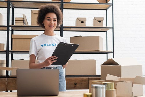 african american volunteer with clipboard smiling at camera near laptop and blurred tins