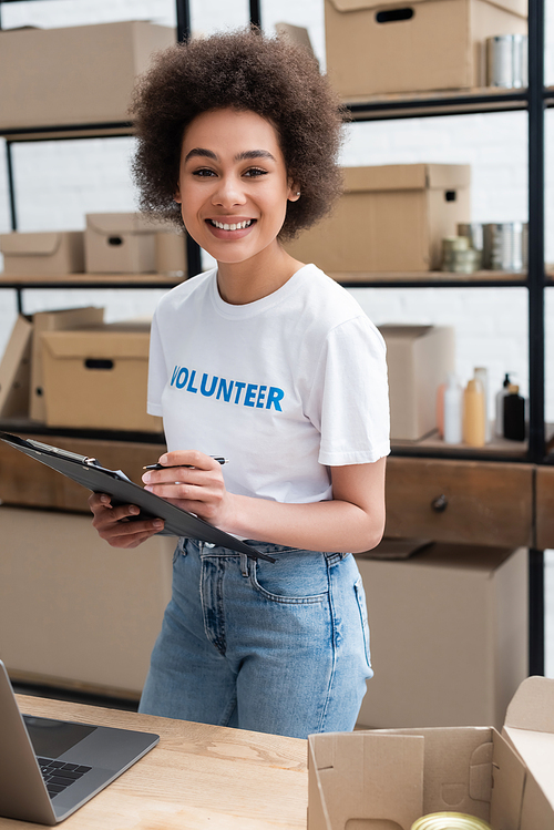 happy african american woman with clipboard and pen smiling at camera in volunteer warehouse