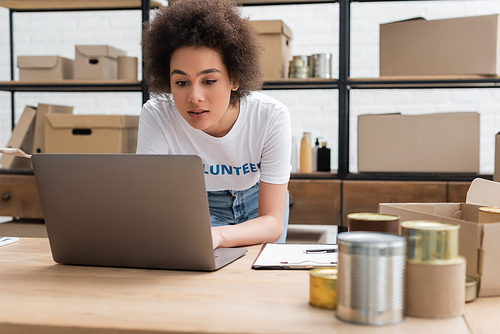 african american volunteer working on laptop near blurred tins in charity center