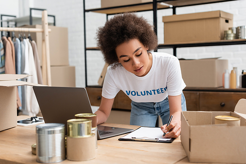 young african american volunteer writing on clipboard near laptop in donation center