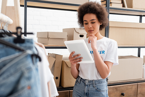 pensive african american volunteer looking at digital tablet near rack with carton boxes