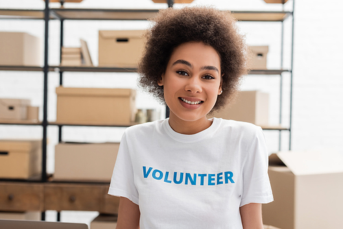 happy african american woman in t-shirt with volunteer lettering looking at camera in charity center