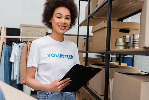 happy african american woman with clipboard near rack with boxes and canned food