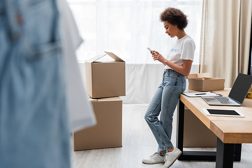 full length of african american volunteer with smartphone near work desk and boxes in charity center