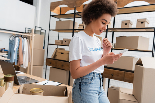 positive african american volunteer using smartphone while working in donation storehouse