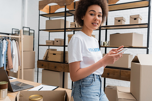 positive african american volunteer using smartphone and looking at camera in charity warehouse