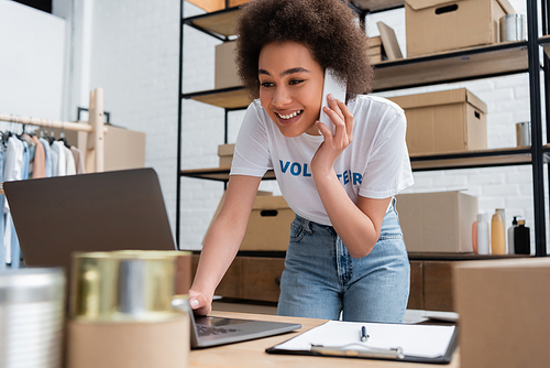 happy african american volunteer talking on smartphone near laptop in donation center