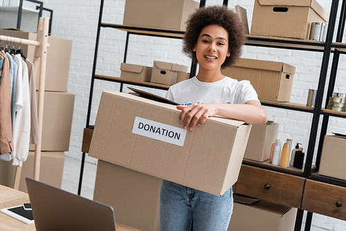 young african american volunteer holding box with donation lettering in warehouse