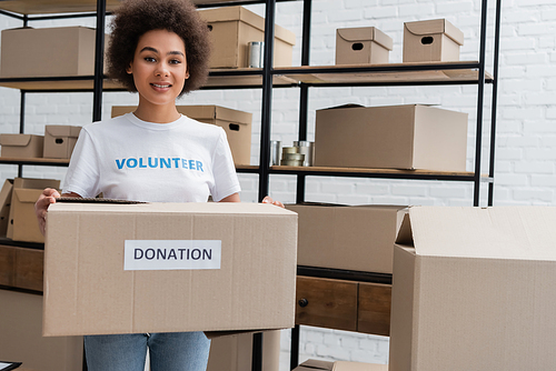 african american volunteer smiling at camera while holding donation box in charity warehouse