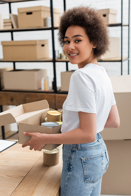 pleased african american volunteer holding box with canned food in charity center