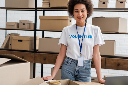 young african american volunteer with name tag smiling at camera near carton boxes