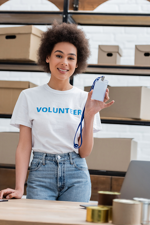 happy african american volunteer showing blank name tag at camera in charity center