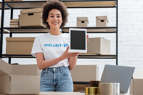 happy african american volunteer showing digital tablet with blank screen in donation warehouse