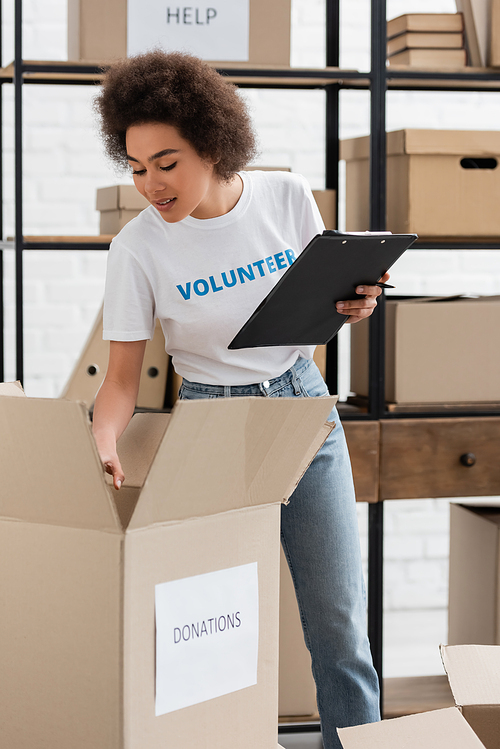 young african american volunteer with clipboard checking carton box in donation warehouse