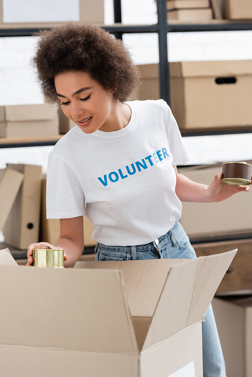 smiling african american volunteer holding canned food near box in charity storehouse