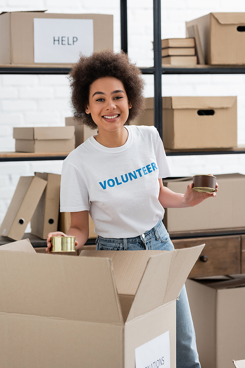 joyful african american volunteer packing canned foodstuff in cardboard box