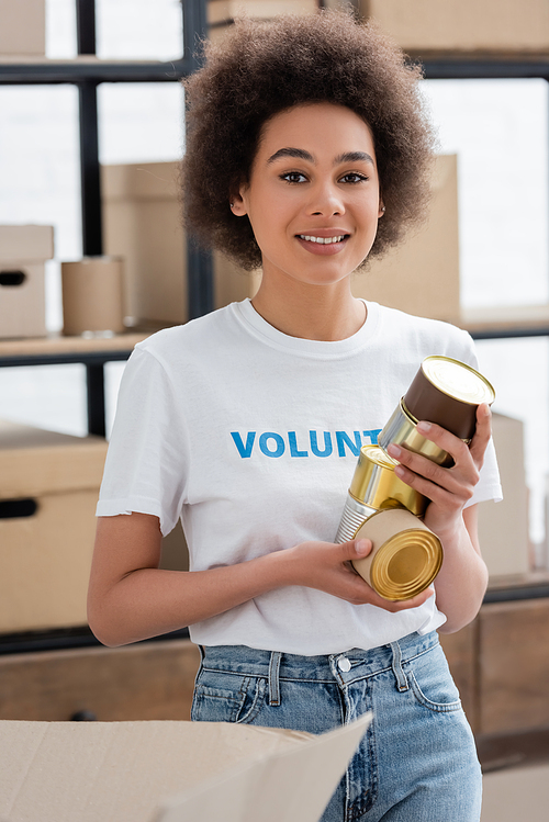 young african american woman with canned food smiling at camera in volunteer center