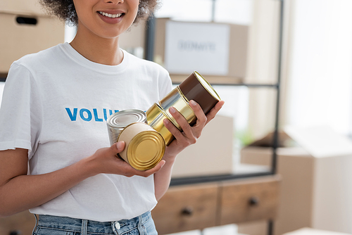 cropped view of smiling african american volunteer holding canned food in charity warehouse