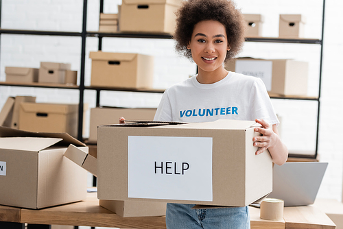 happy african american volunteer holding carton box with help lettering in charity center