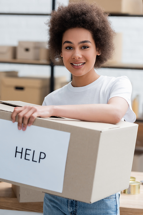 young african american volunteer smiling at camera near box with help lettering