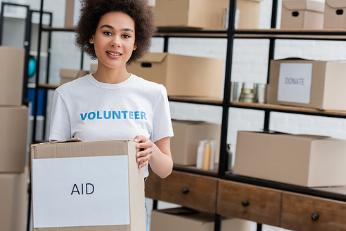 smiling african american volunteer holding box with aid lettering in charity storehouse