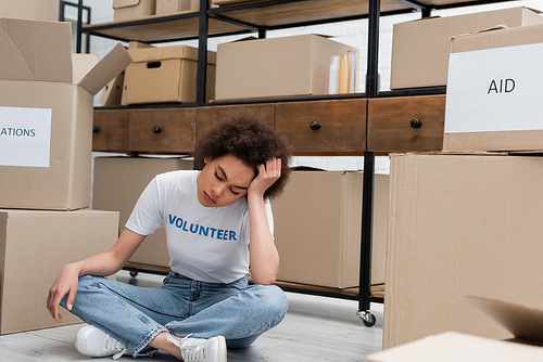 tired african american volunteer sitting on floor near rack with donation boxes