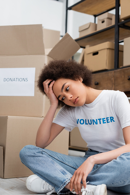 tired african american woman sitting near cardboard boxes in charity center