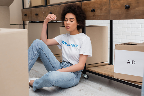 exhausted african american volunteer with closed eyes sitting on floor in charity warehouse