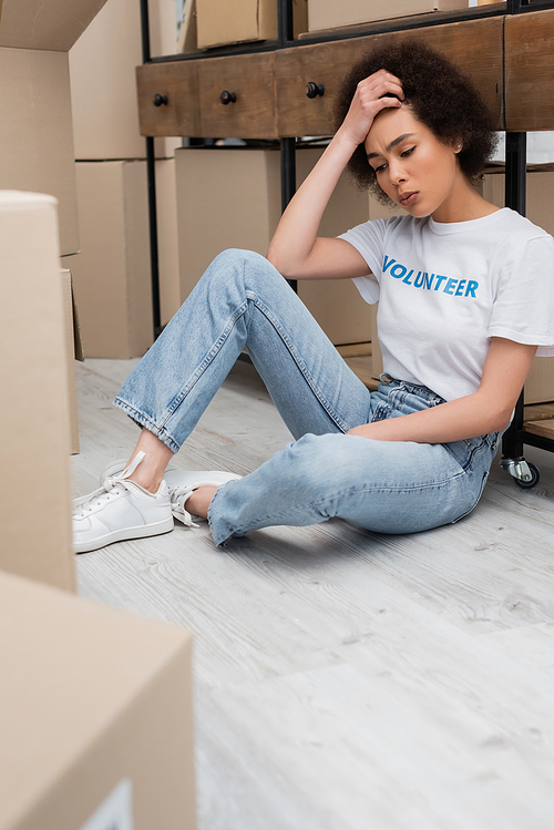 exhausted african american volunteer sitting on floor near carton boxes in charity center