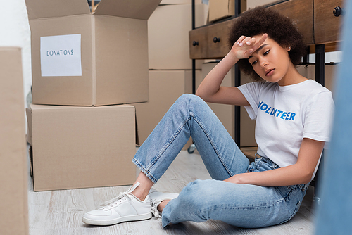 tired african american woman sitting on floor near donation boxes in charity center