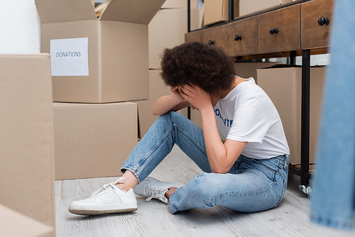 exhausted african american woman obscuring face with hands while sitting on floor in volunteer center