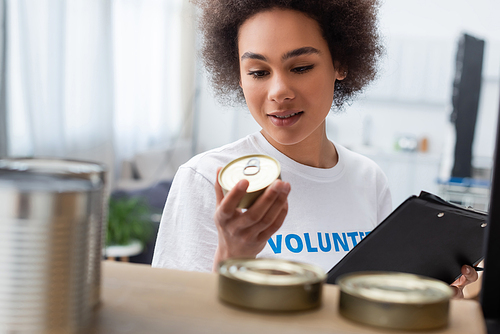 smiling african american woman with clipboard holding canned food in charity center